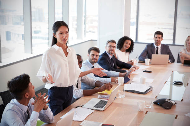 a woman in white formal shirt standing up while talking during a work meeting while her coworkers listens